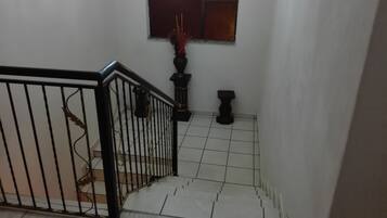 Una escalera con barandilla de metal negro, suelo de baldosas blancas y un pequeño ventanal con un adorno rojo.