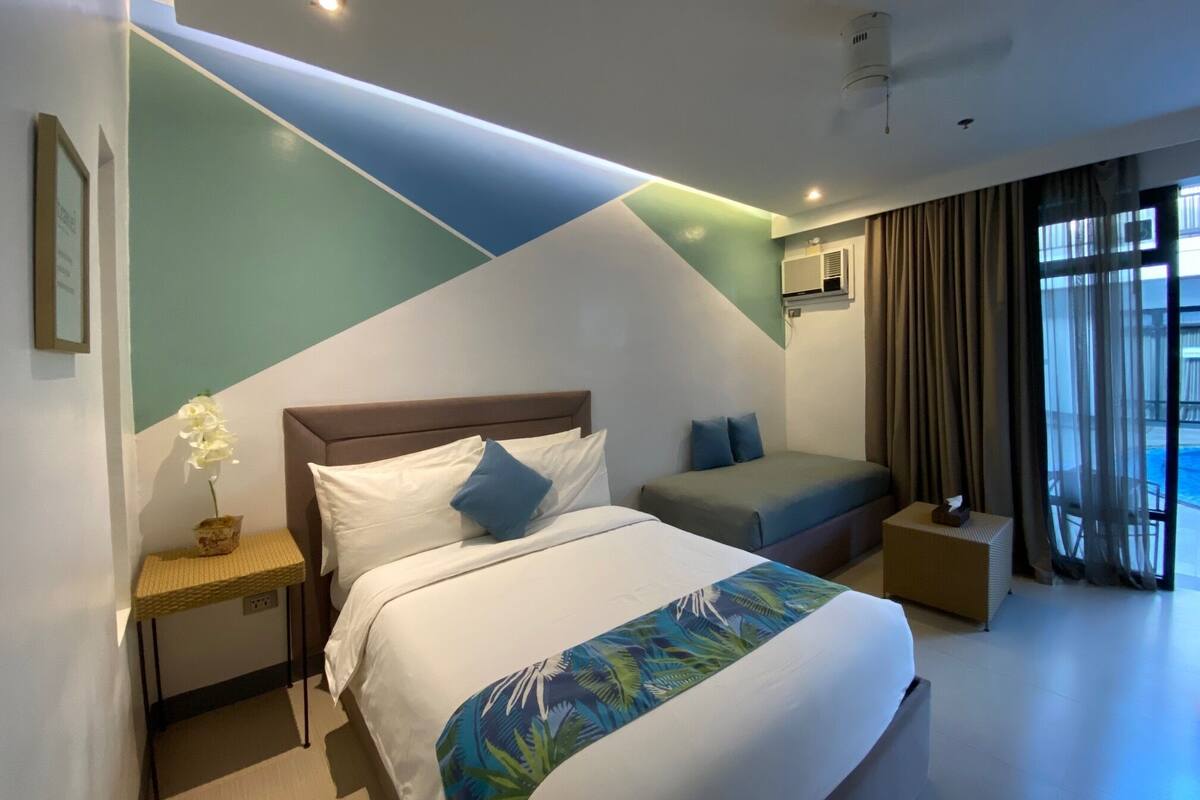 A modern hotel room with a large bed, a sofa, a small table, and a view of the pool.