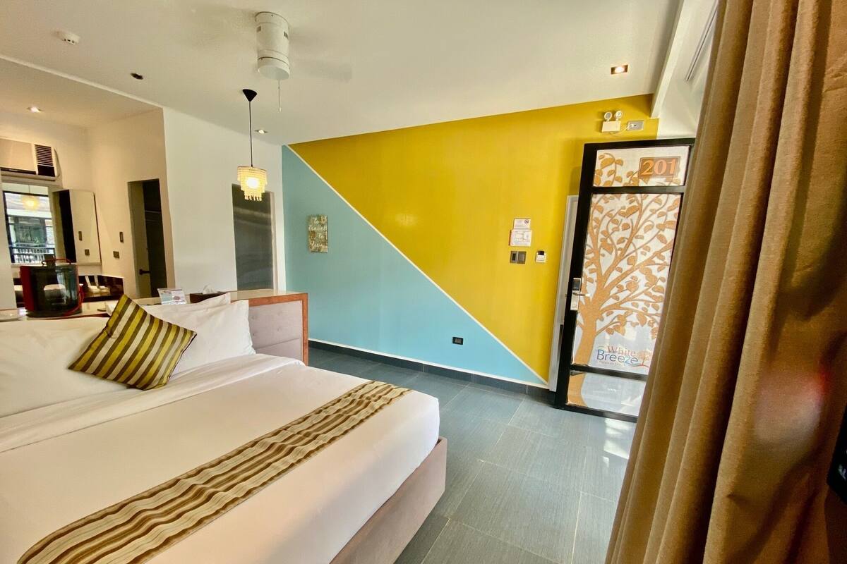 A hotel room with a bed, a bedside table, a mirror, a wall with a yellow and blue design, and a door with a patterned design.
