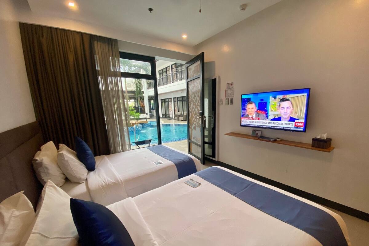 A hotel room with two beds, a TV on the wall, and a view of an outdoor swimming pool.