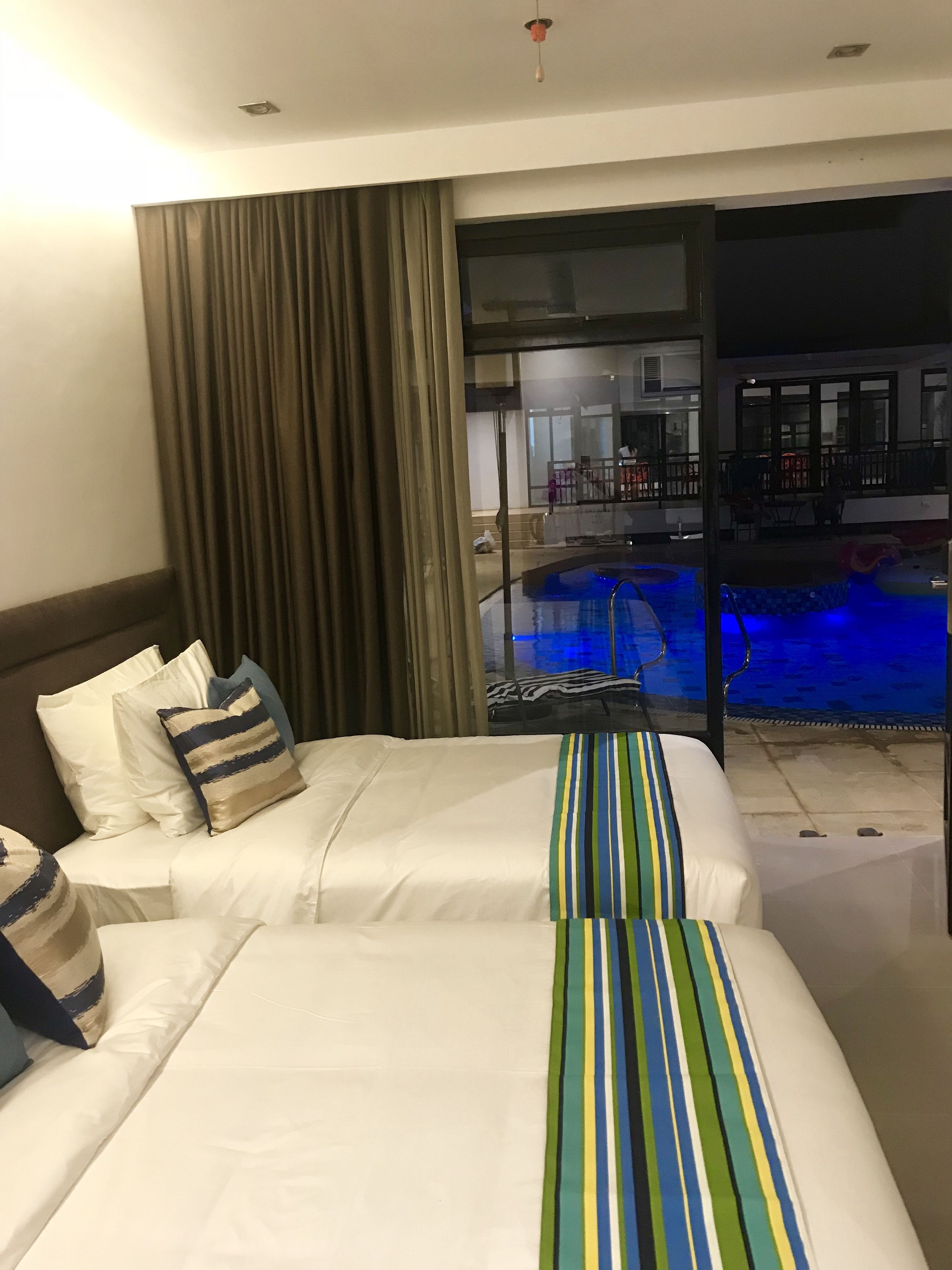 A hotel room with two beds, a large window with curtains, a view of a swimming pool, and a balcony.
