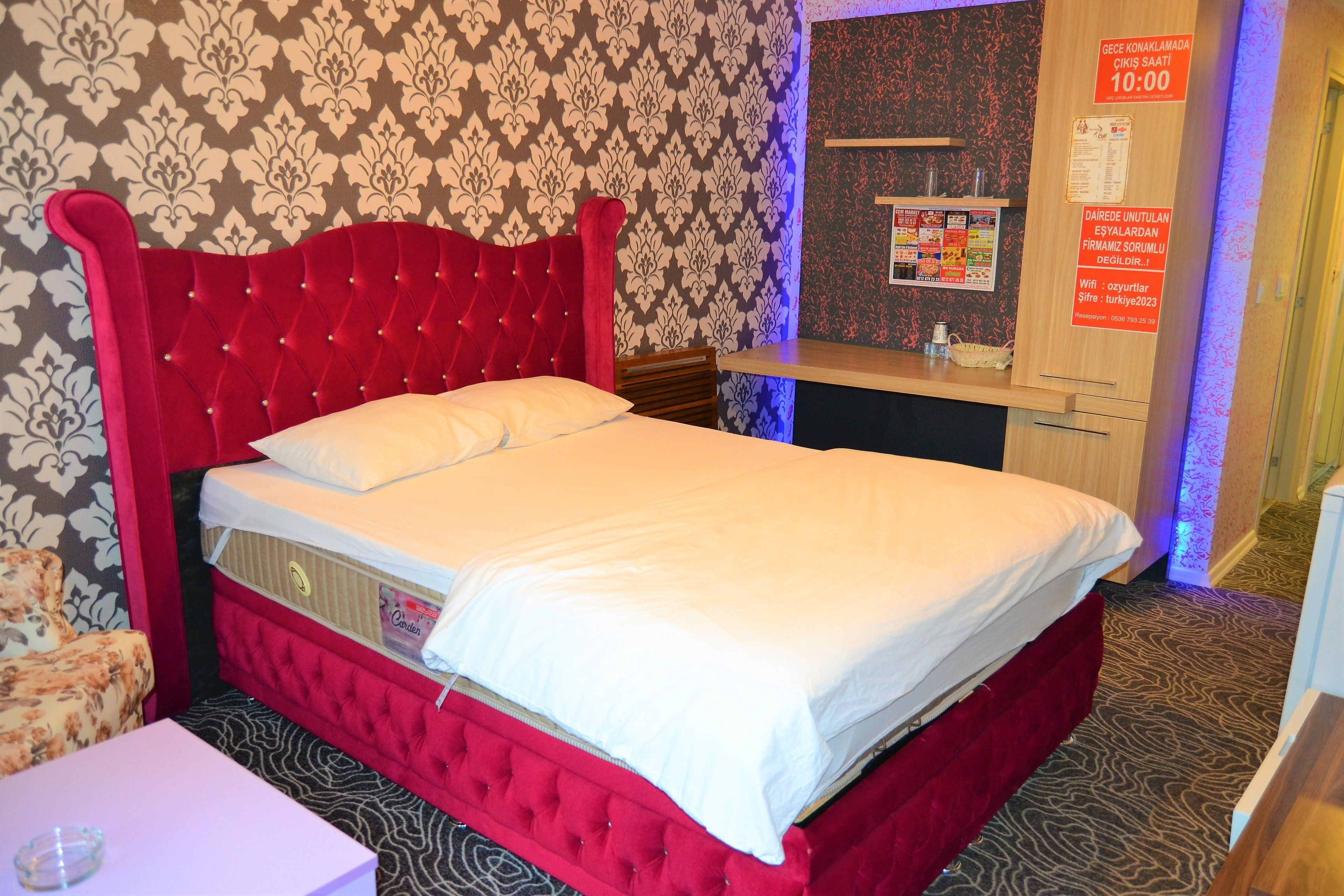 A hotel room with a red upholstered bed, a white pillow, a wooden nightstand, and a mirror.