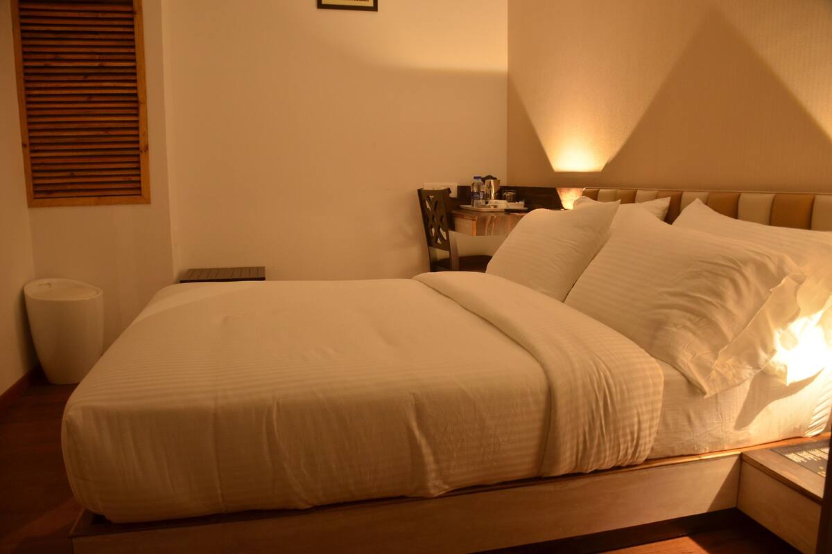 A bed with a headboard, a bedside table with a lamp, and a window with blinds.