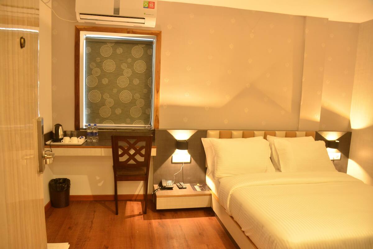 A hotel room with a bed, a desk with a chair, a window with patterned curtains, and an air conditioning unit.