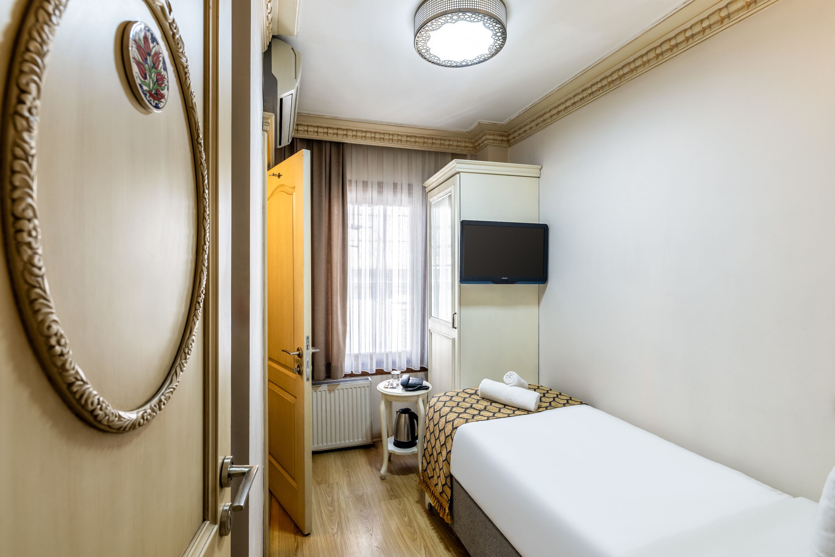Habitación de hotel con una cama, una mesita con hervidor, un radiador, una ventana con cortinas y un televisor en la pared.