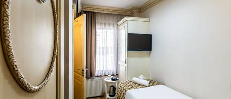 Habitación de hotel con una cama, una mesita con hervidor, un radiador, una ventana con cortinas y un televisor en la pared.