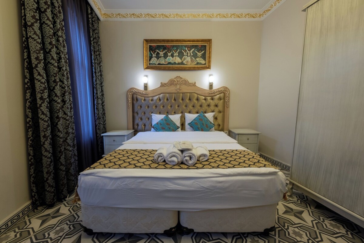 A hotel room with a large bed, a headboard with lights, a painting on the wall, and patterned curtains.