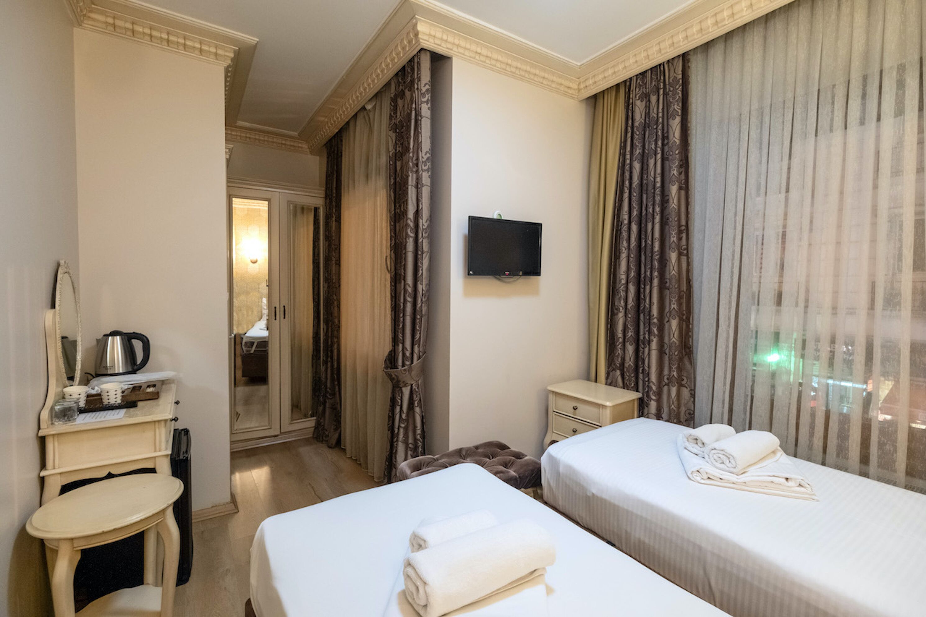 A hotel room with two beds, a desk, a chair, a television, and a view of the city through the window.