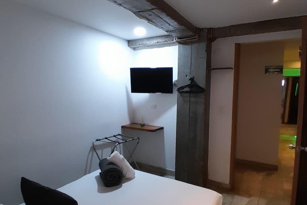 A bedroom with a bed, a TV mounted on the wall, a small table, and a door leading to another room.