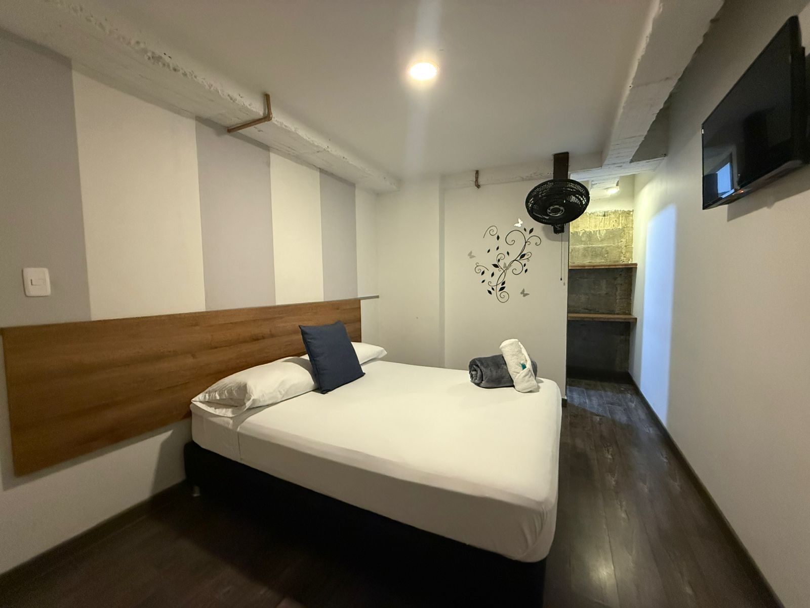Un dormitorio con cabecera de madera, cama con ropa de cama blanca, mesita de noche pequeña, televisor montado en la pared y un adorno en la pared.