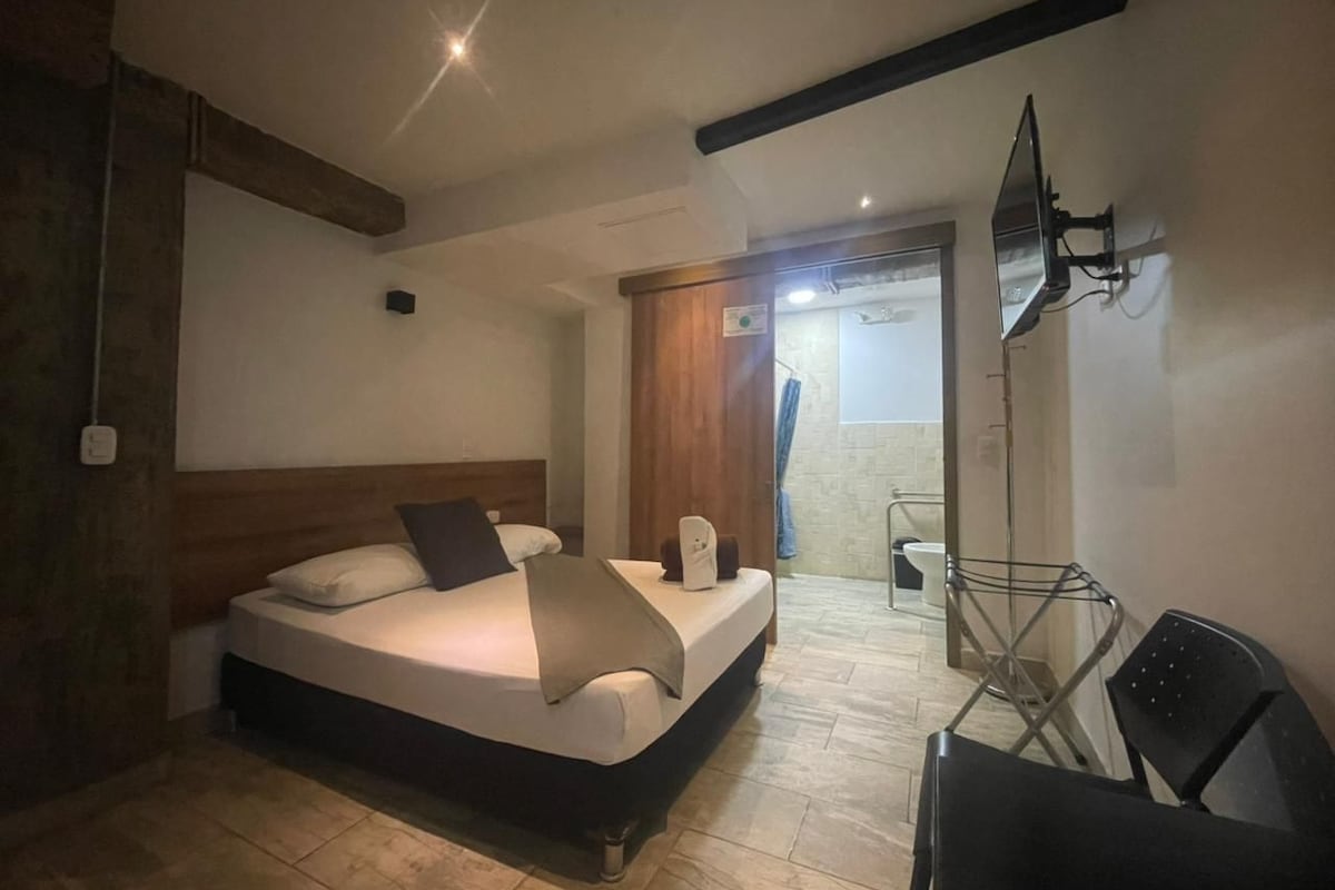A bedroom with a bed, a chair, a TV, and a bathroom visible in the background.