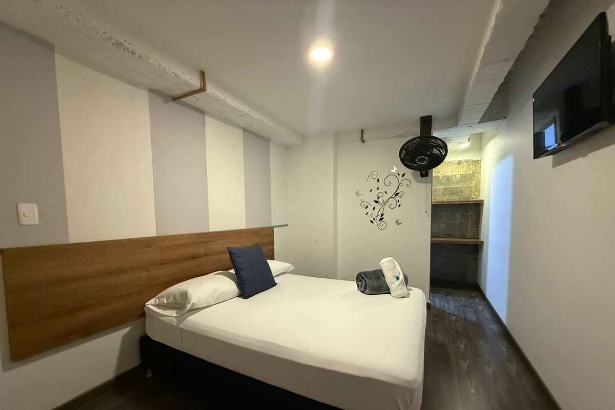 A bedroom with a wooden headboard, a bed with white bedding, a small bedside table, a wall-mounted TV, and a decorative wall piece.