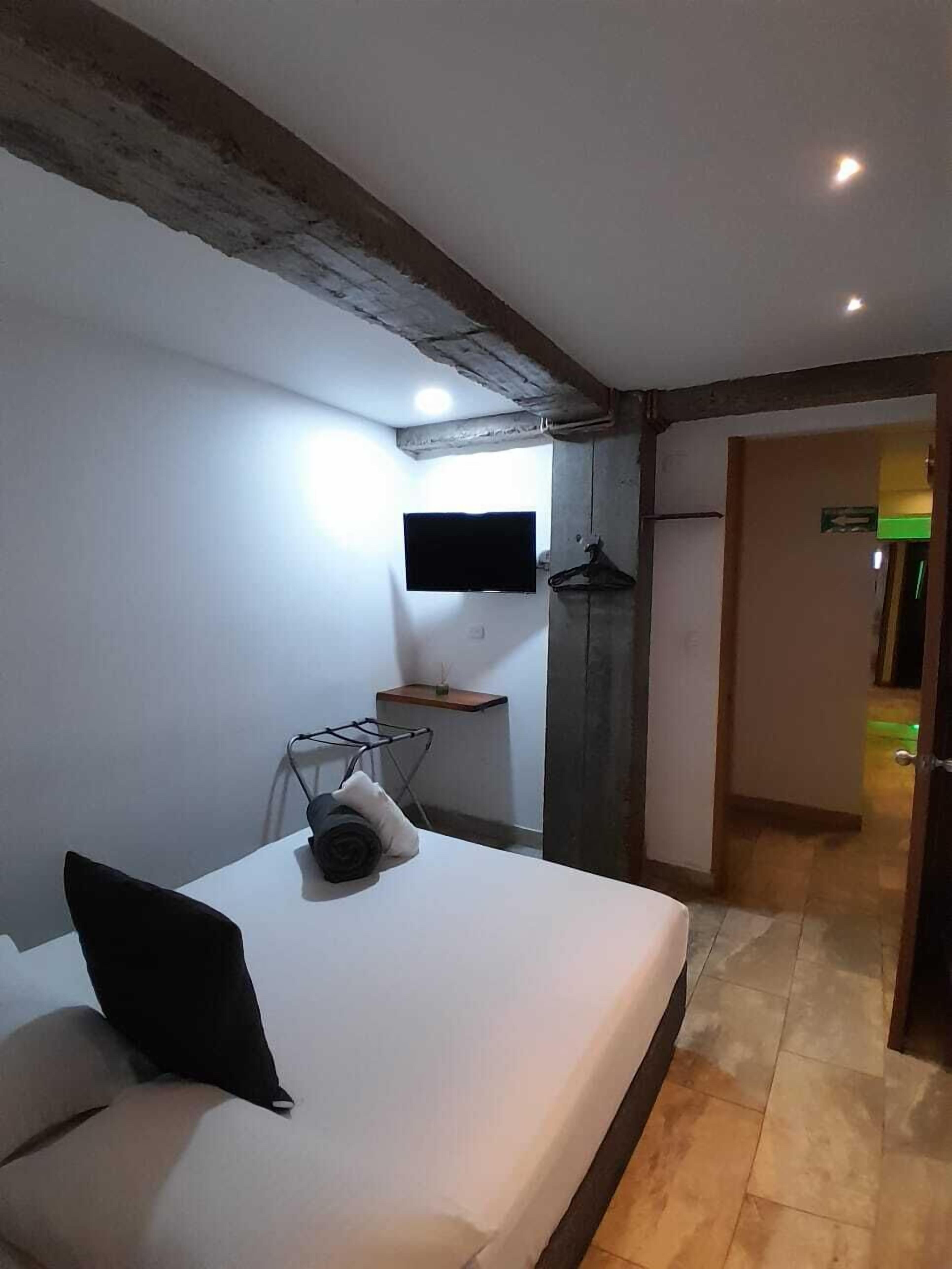 Un dormitorio con una cama, un televisor montado en la pared, una mesita y una puerta que da a otra habitación.