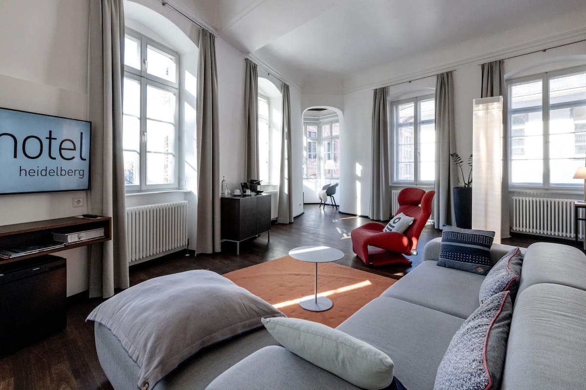 A modern hotel room with a large sofa, a red armchair, a flat-screen TV, and a view of the city through large windows.