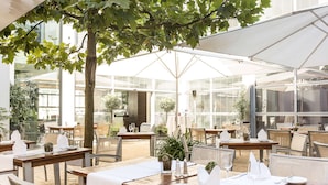 An outdoor dining area with tables and chairs, covered by umbrellas, and surrounded by glass walls. - Arthotel Heidelberg (Heidelberg)