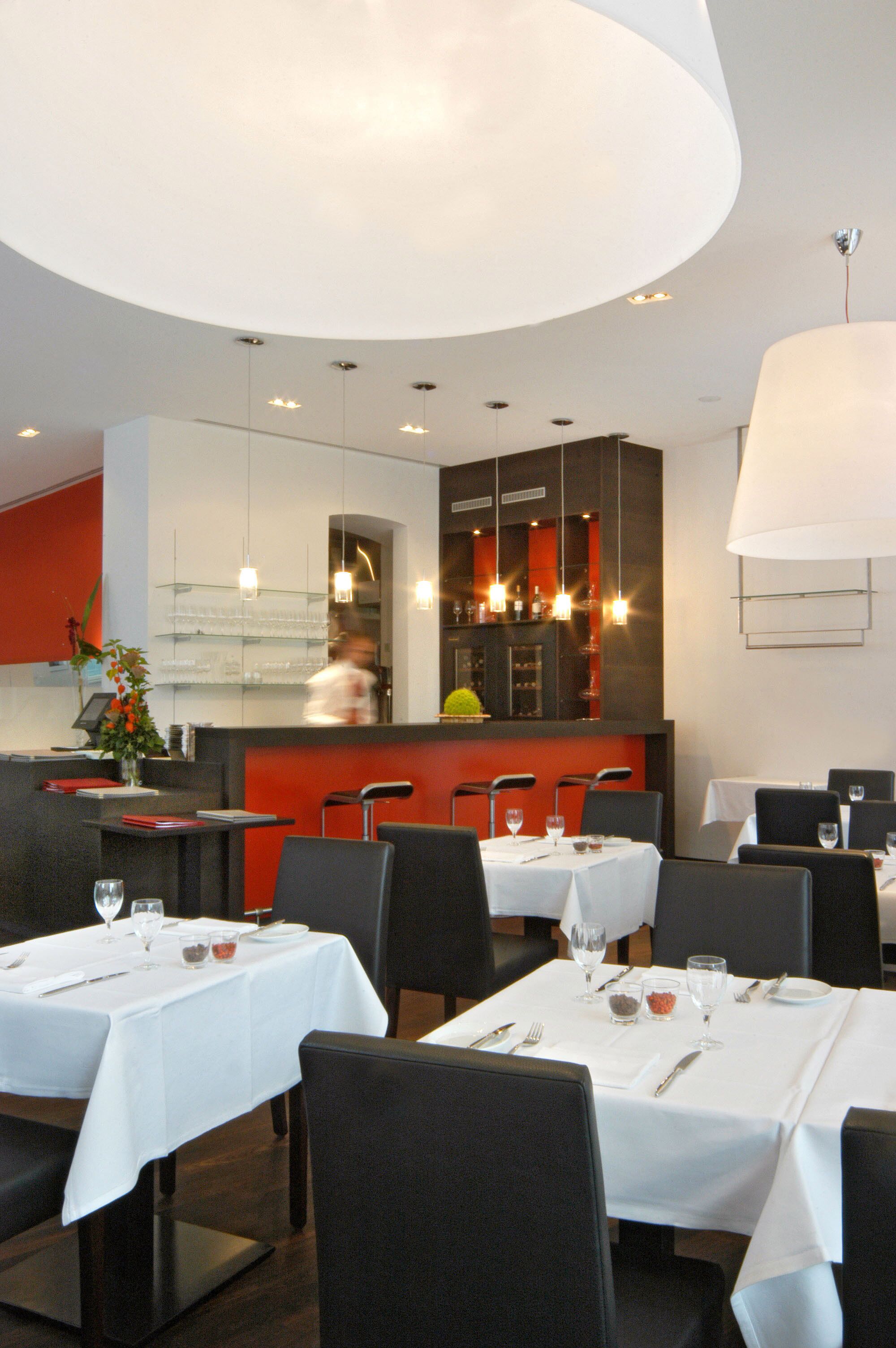 Un restaurante con mesas dispuestas para comer, una zona de bar y un chef al fondo.