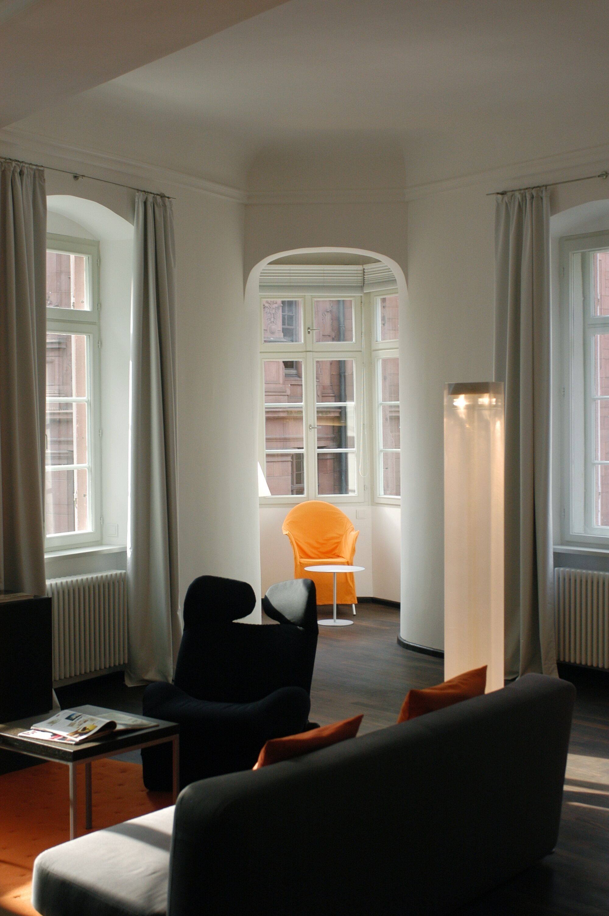 Habitación de hotel con diseño moderno, que incluye un ventanal grande, un sillón negro, una silla pequeña naranja y una lámpara de pie.