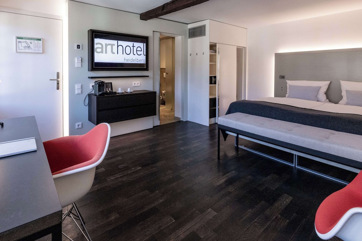 A modern hotel room with a bed, a desk, a chair, a TV, and a wardrobe.