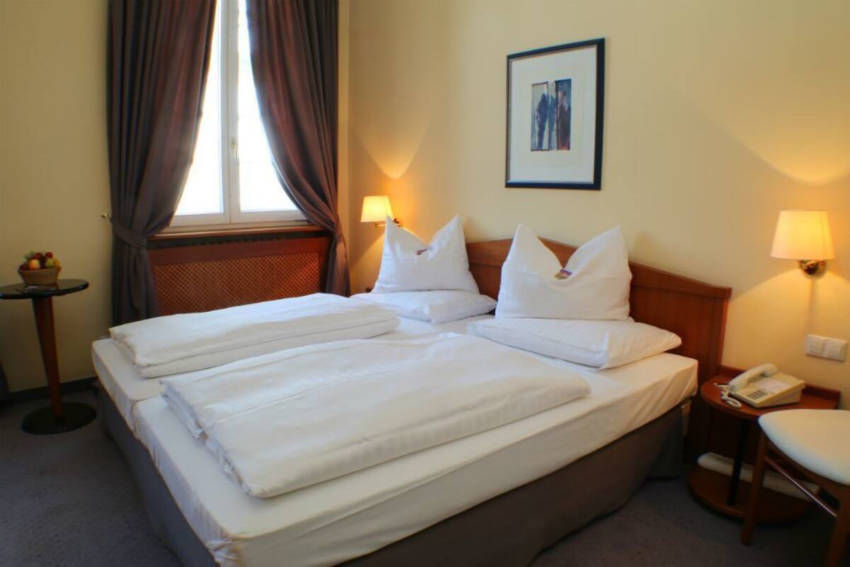 Une chambre d’hôtel avec un lit double, deux tables de chevet, une lampe, un tableau encadré au mur et une fenêtre avec des rideaux.