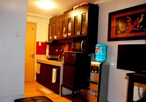 Una cocina compacta con armarios de madera, un refrigerador, un dispensador de agua y un televisor montado en la pared.