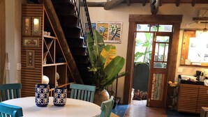 A dining area with a round table, teal chairs, and patterned vases. A staircase with wooden railings leads to a kitchen area. - Casa Carmel (Búzios)