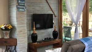 A cozy living area with a stone wall, a flat-screen TV, wooden furniture, and a sign that reads "the Beach is my Happy Place". - Casa Carmel (Búzios)