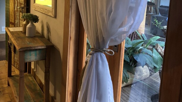 A room with a wooden floor, a wooden table with a vase, a mirror on the wall, and a curtain tied at the bottom. - Casa Carmel (Búzios)