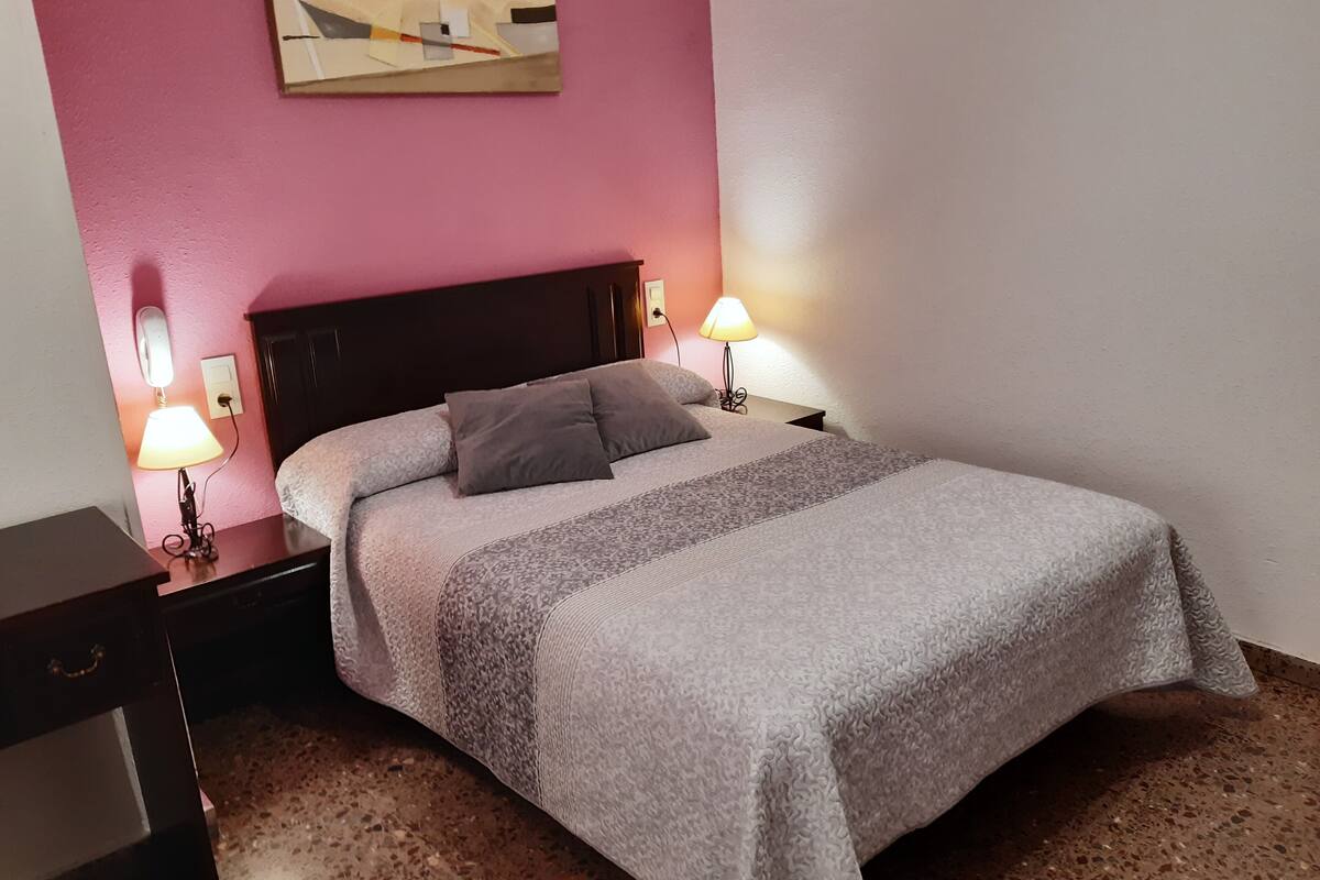 Un lit bien fait, recouvert d’une couvre-lit à motifs, avec deux lampes de chevet et un petit tableau accroché au mur.