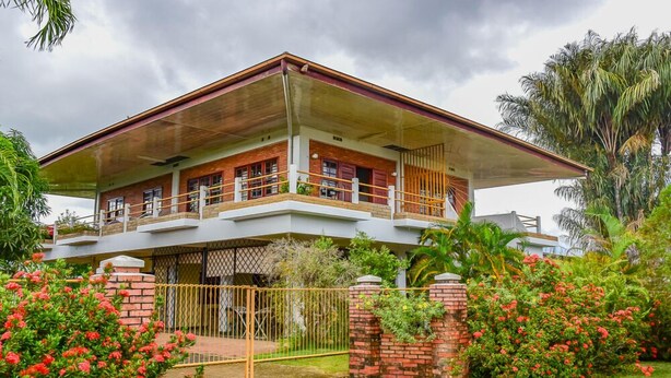 A two-story house with a balcony, surrounded by lush greenery and a gated entrance. - Macsapartment (Paramaribo)