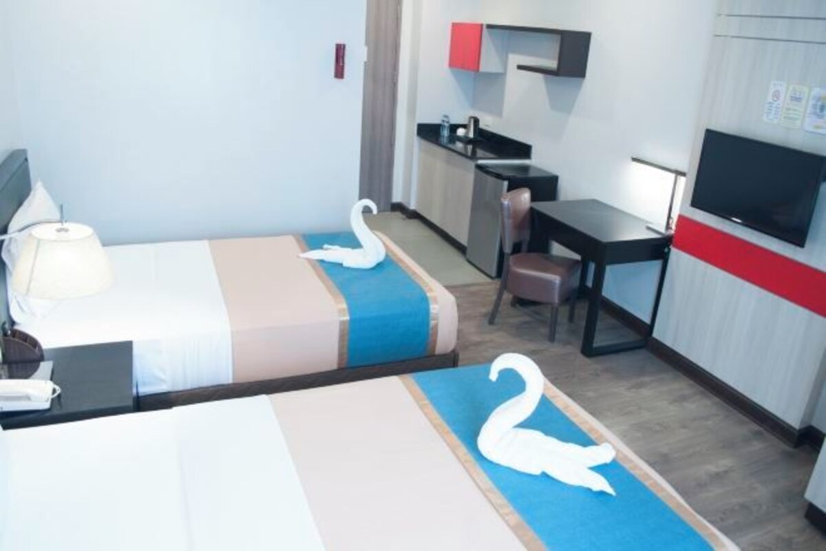 A hotel room with two beds, a desk, a chair, a TV, and a kitchenette.