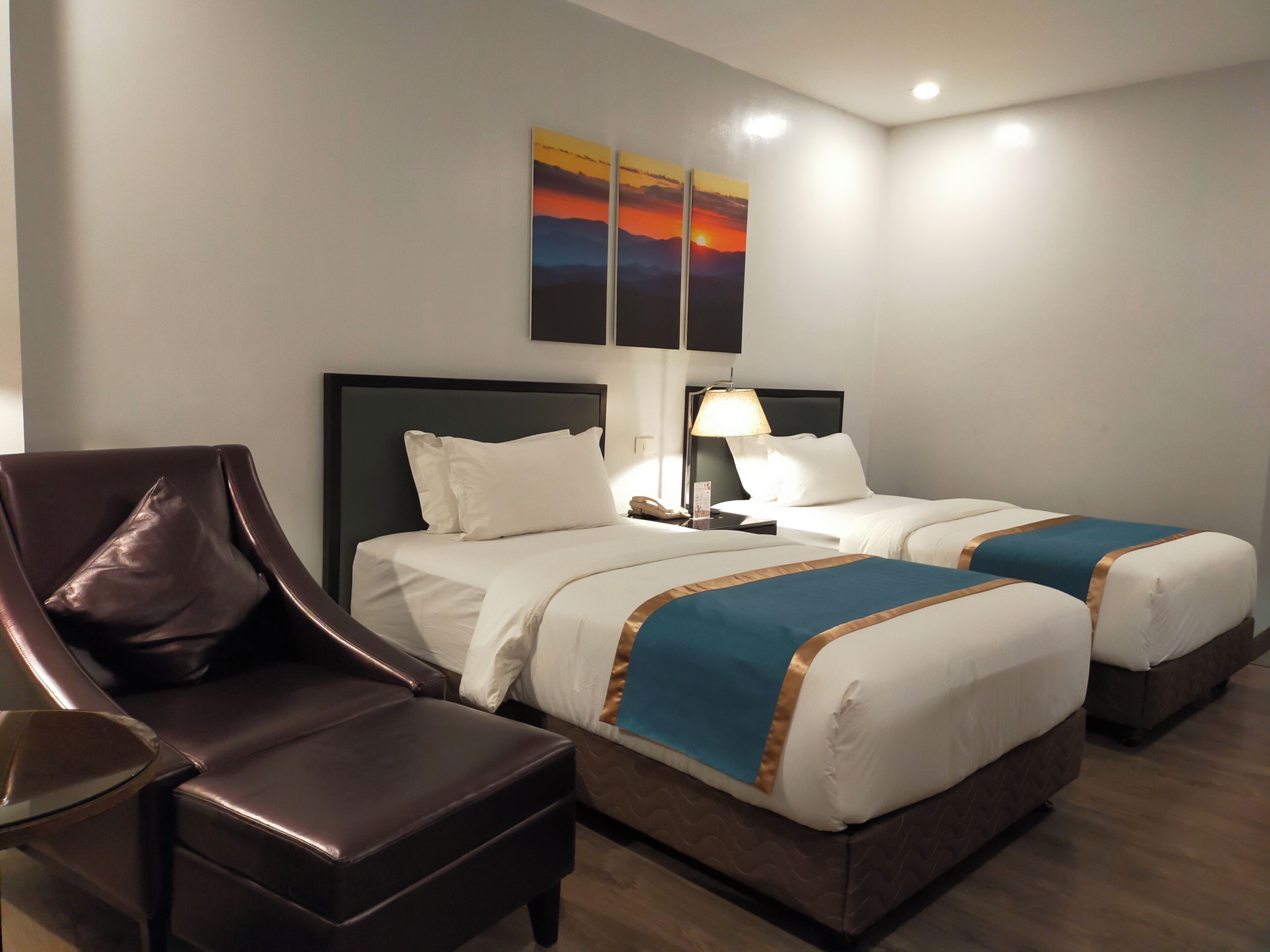 A hotel room with two beds, a chair, a nightstand, and a wall-mounted lamp.
