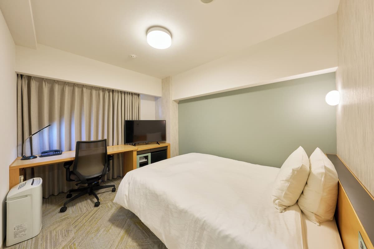 A hotel room with a bed, a desk with a computer and a TV, and a waste bin.