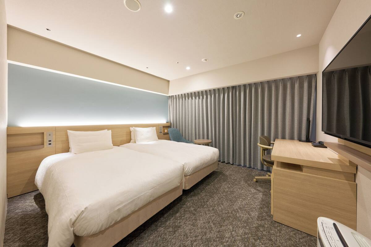 A hotel room with two beds, a desk, a TV, and a large window with curtains.