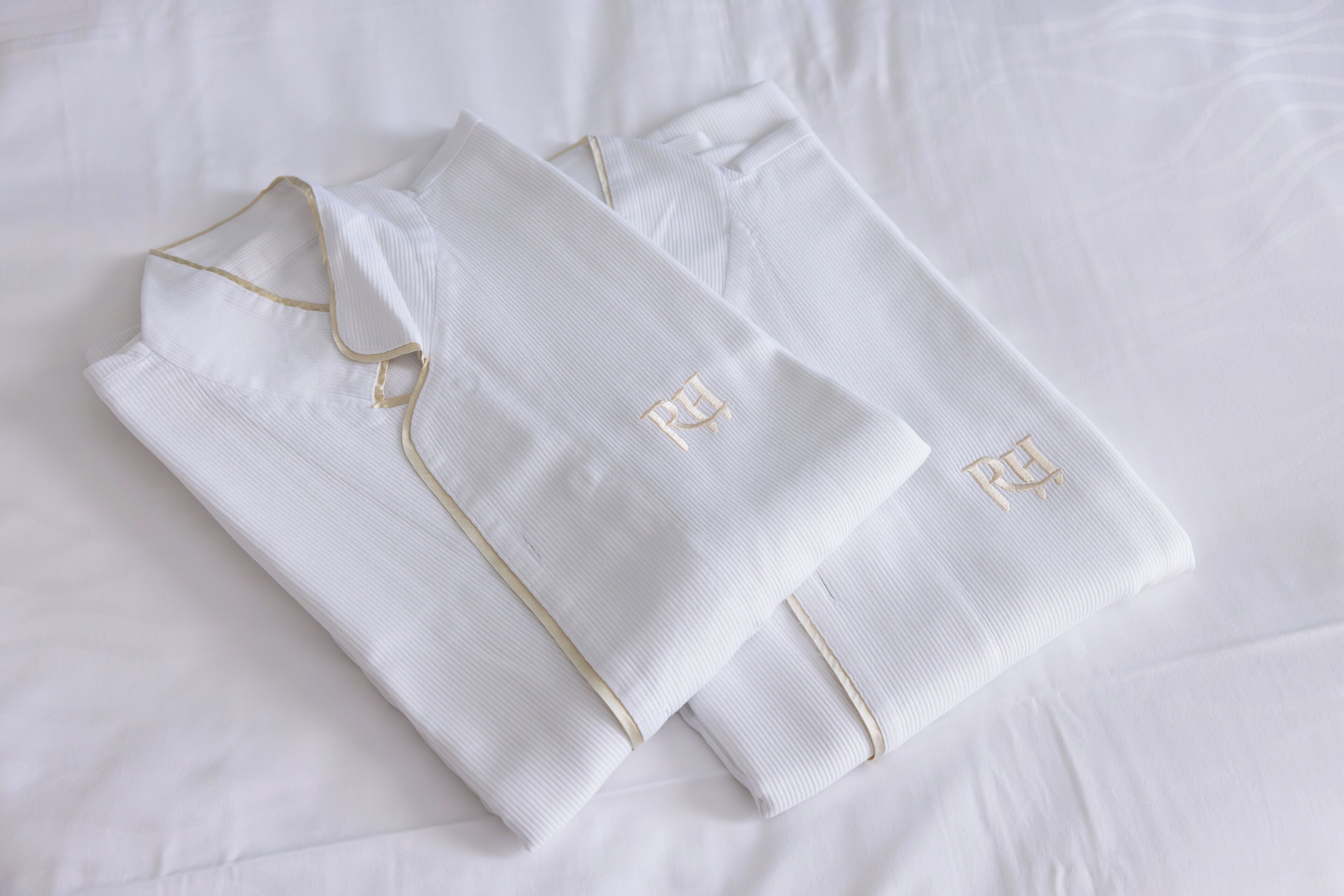 Neatly folded white pajamas with gold trim and embroidered initials on a white bedspread.