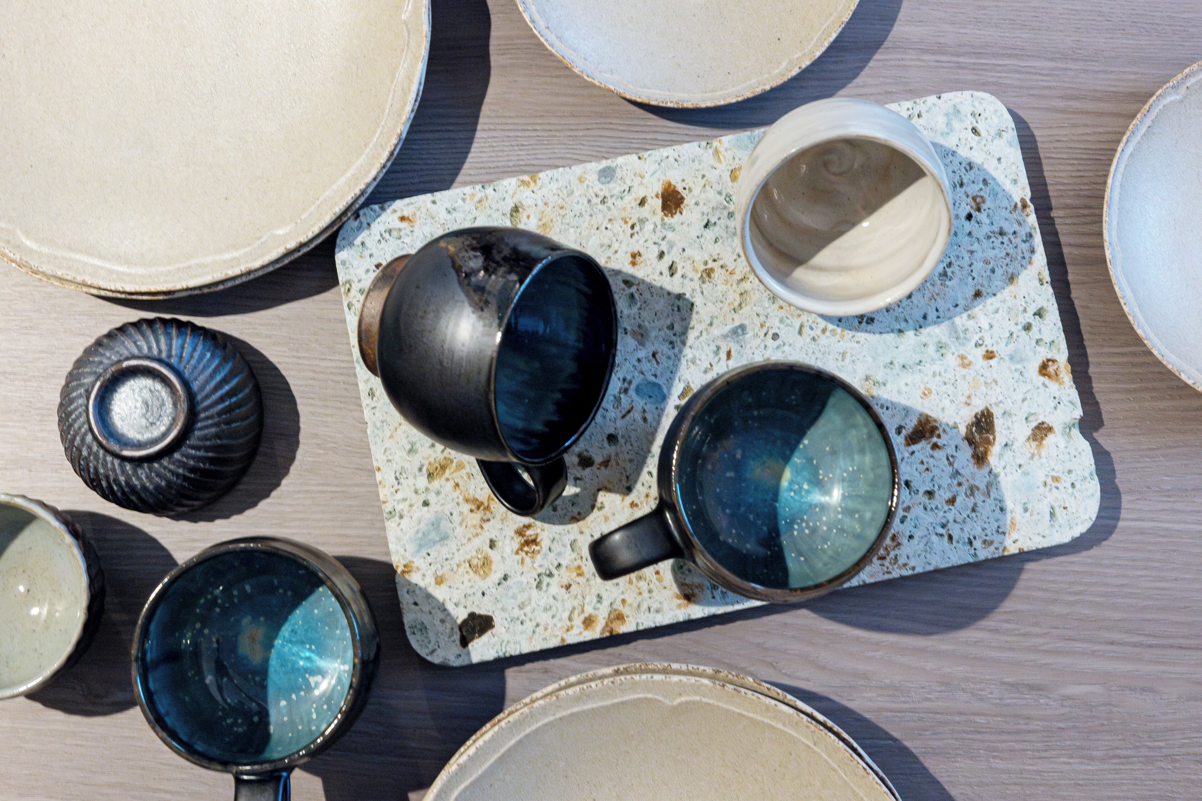 A table with various ceramic dishes and cups, some with reflective surfaces, arranged on a speckled tray.