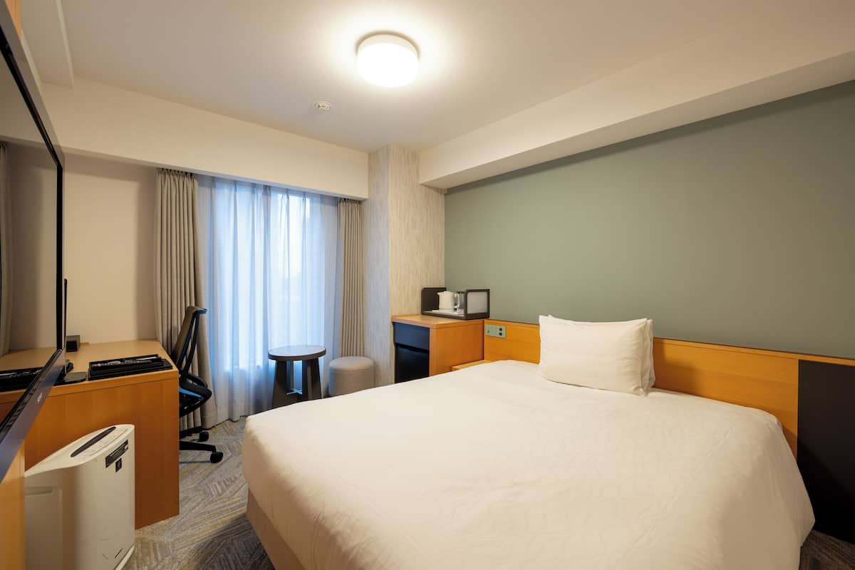 A hotel room with a large bed, a desk with a computer, a chair, a small table, and a window with curtains.