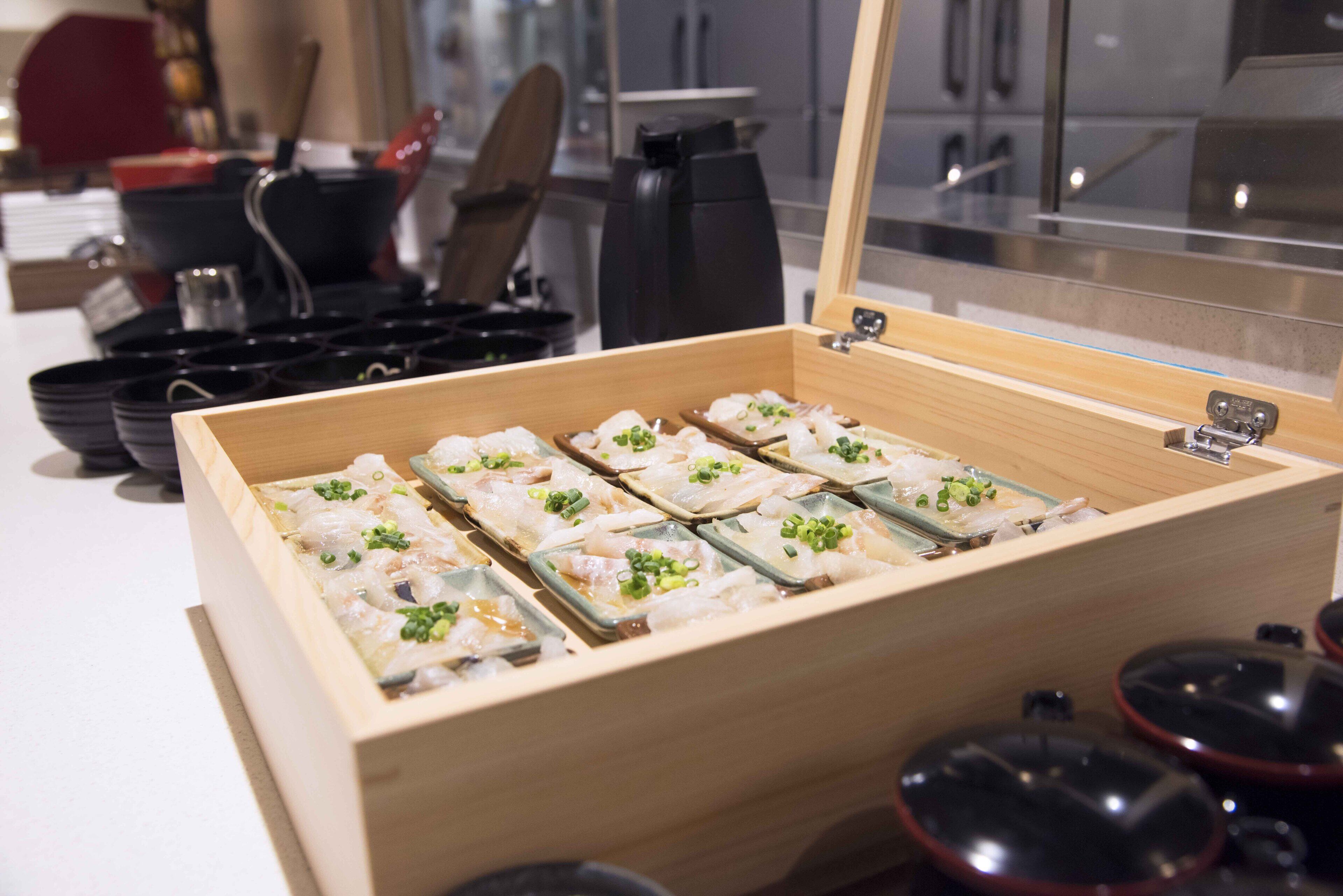 A wooden box with compartments containing seafood dishes, a coffee machine, and black cups in the background.