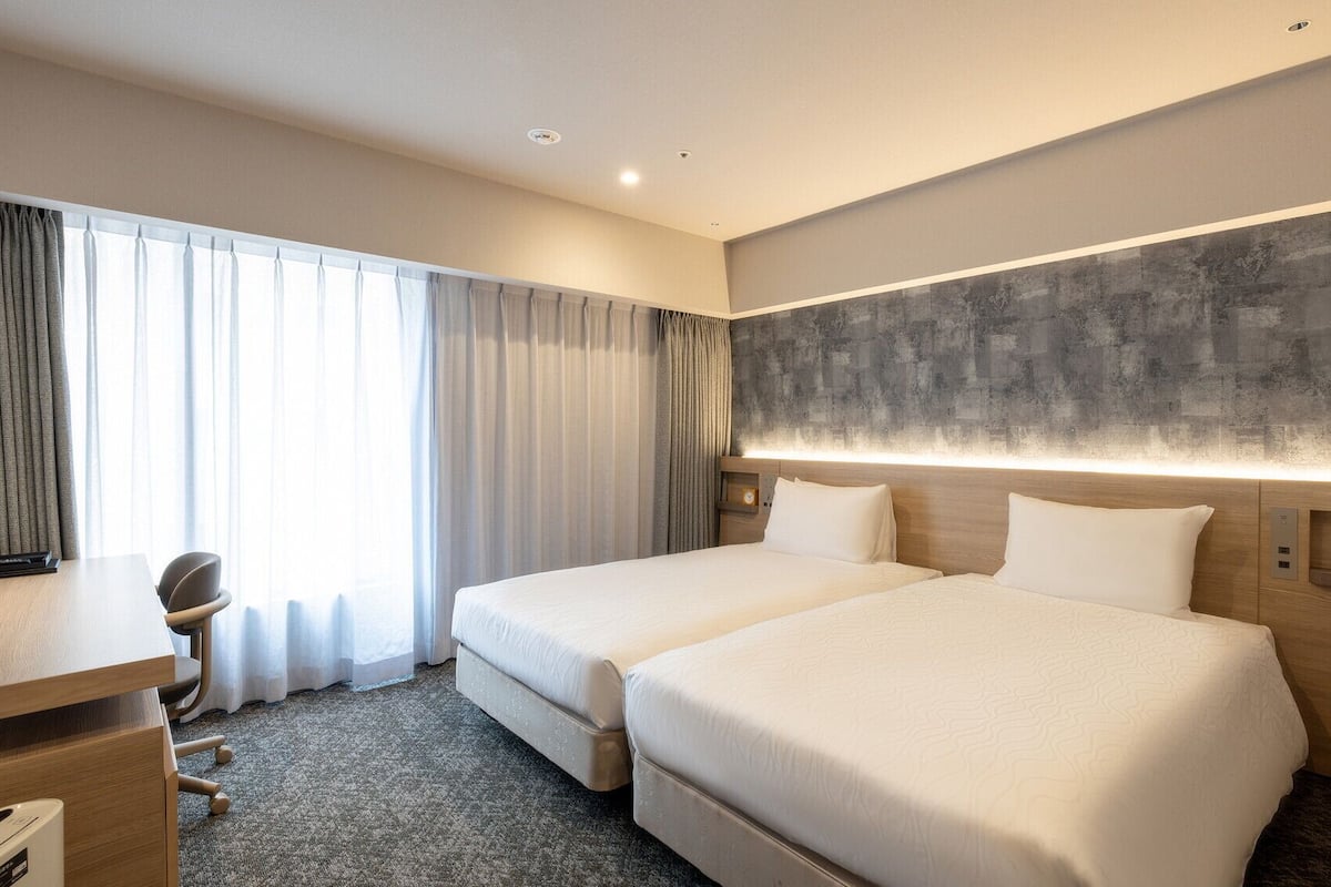 Une chambre d’hôtel avec deux lits, un bureau, une chaise et une grande fenêtre avec des rideaux.