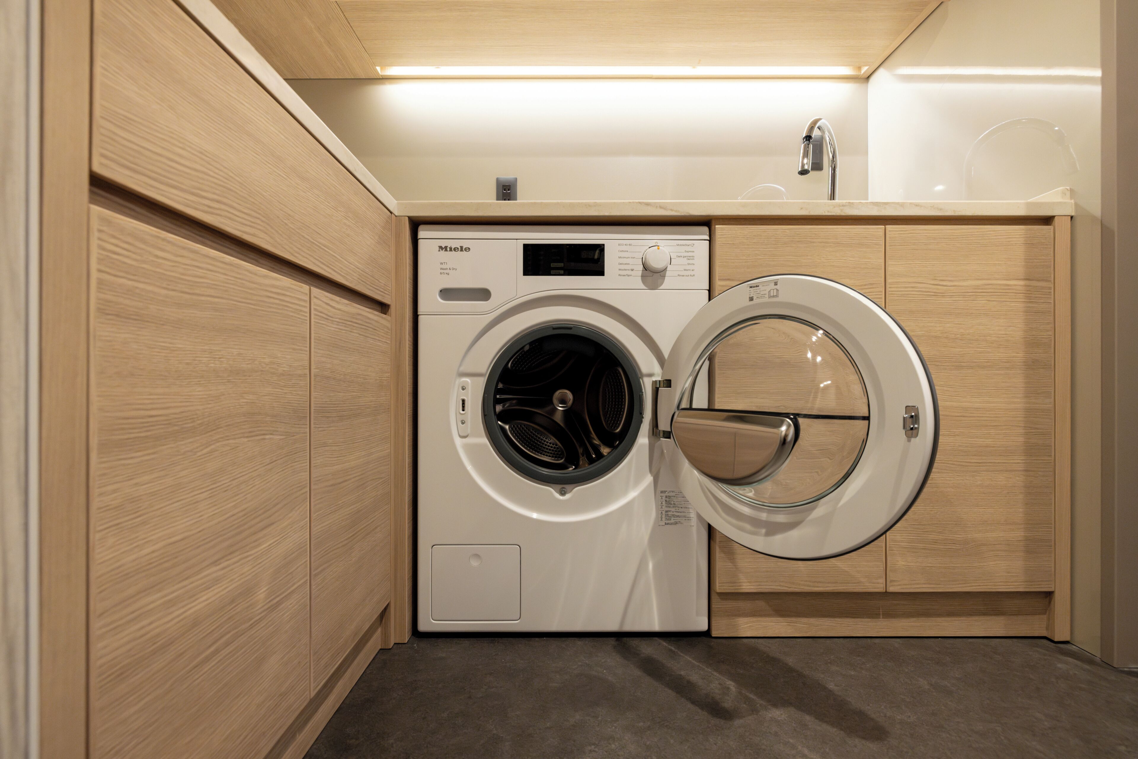 A compact, modern laundry room with a front-loading washing machine, wooden cabinetry, and a countertop with a sink.