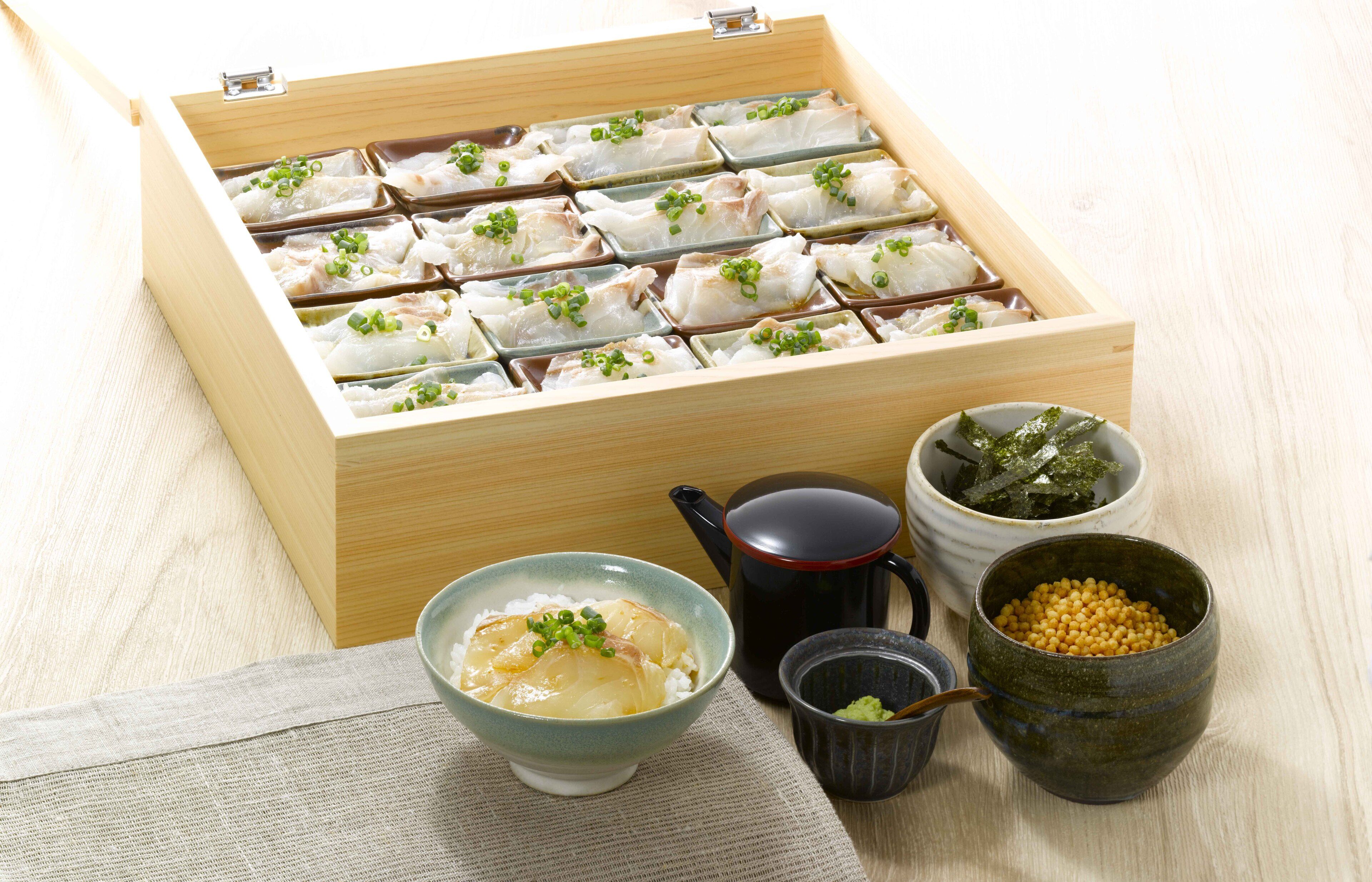 A wooden box with compartments containing food items, a bowl of soup, and two small cups with green and yellow contents.