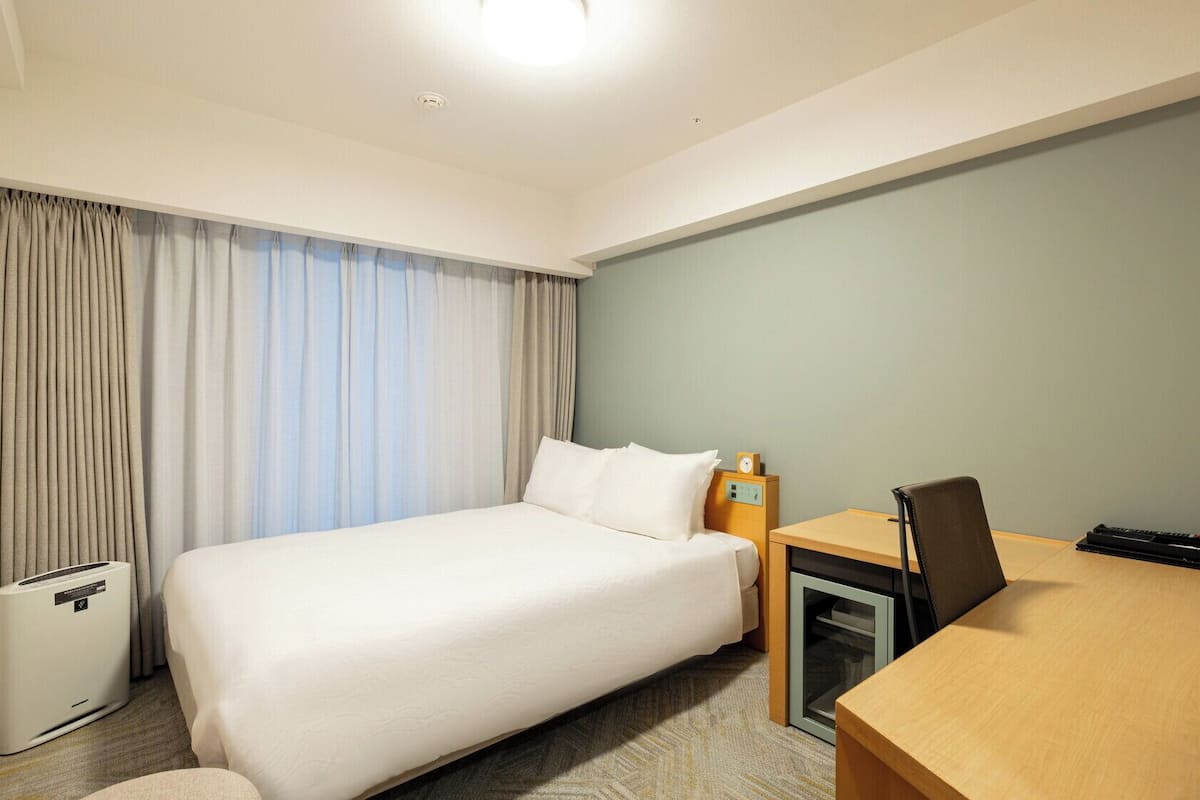 A hotel room with a bed, desk, chair, and a small table with a lamp.