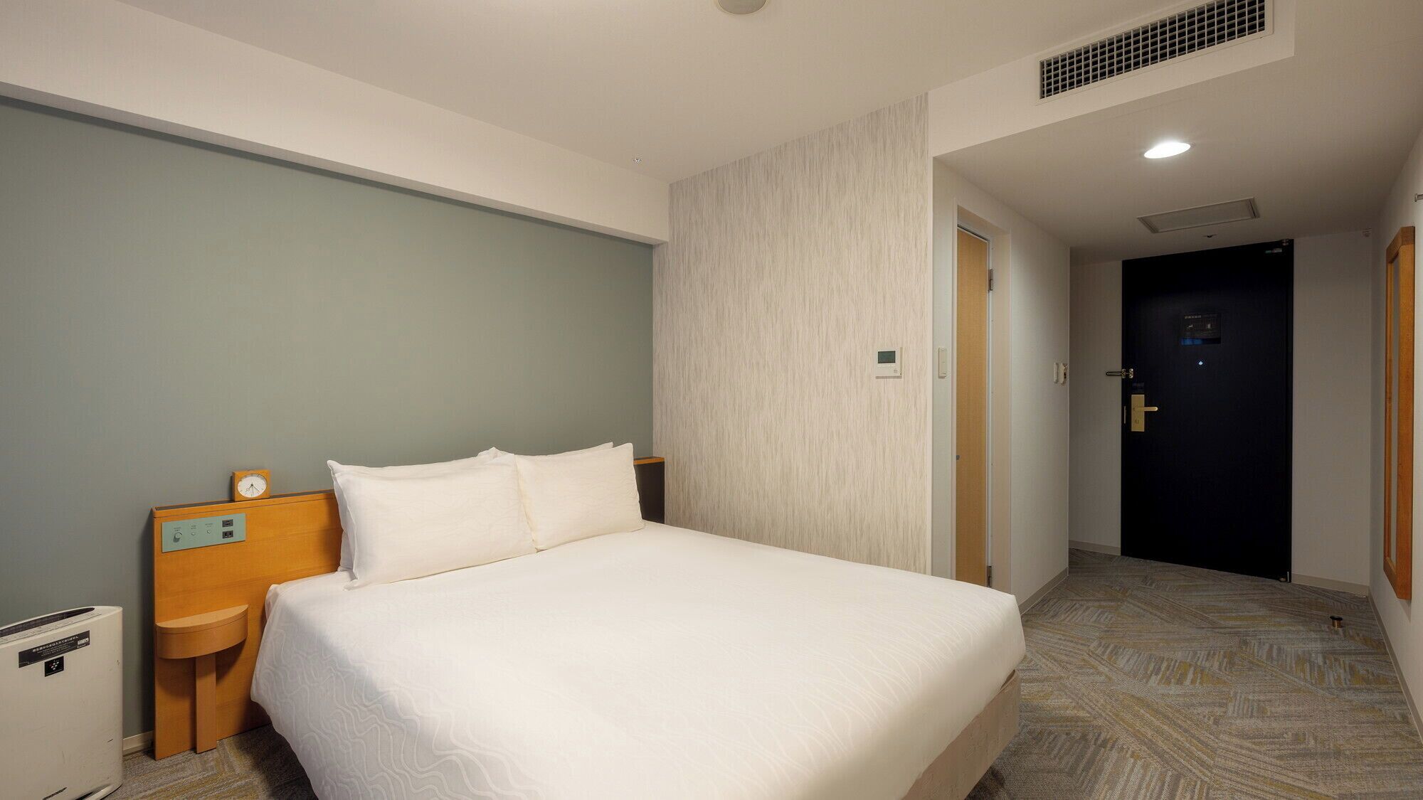 Une chambre d’hôtel comprenant un lit, une table de chevet avec une horloge, un panneau de commande fixé au mur et une porte donnant sur une autre pièce.