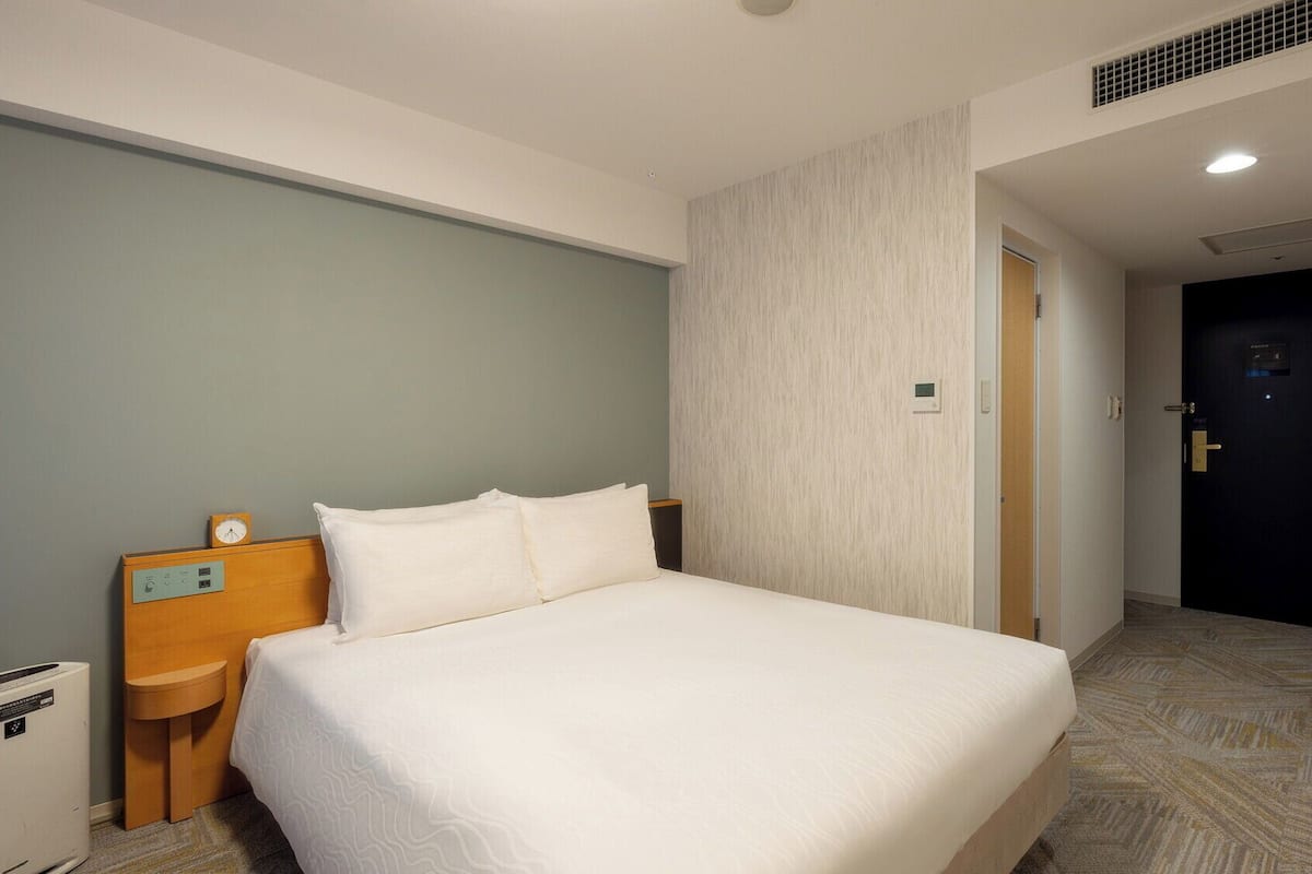 Ein Hotelzimmer mit einem Bett, einem Nachttisch mit einer Uhr, einer Wandbedienung und einer Tür zu einem anderen Raum.