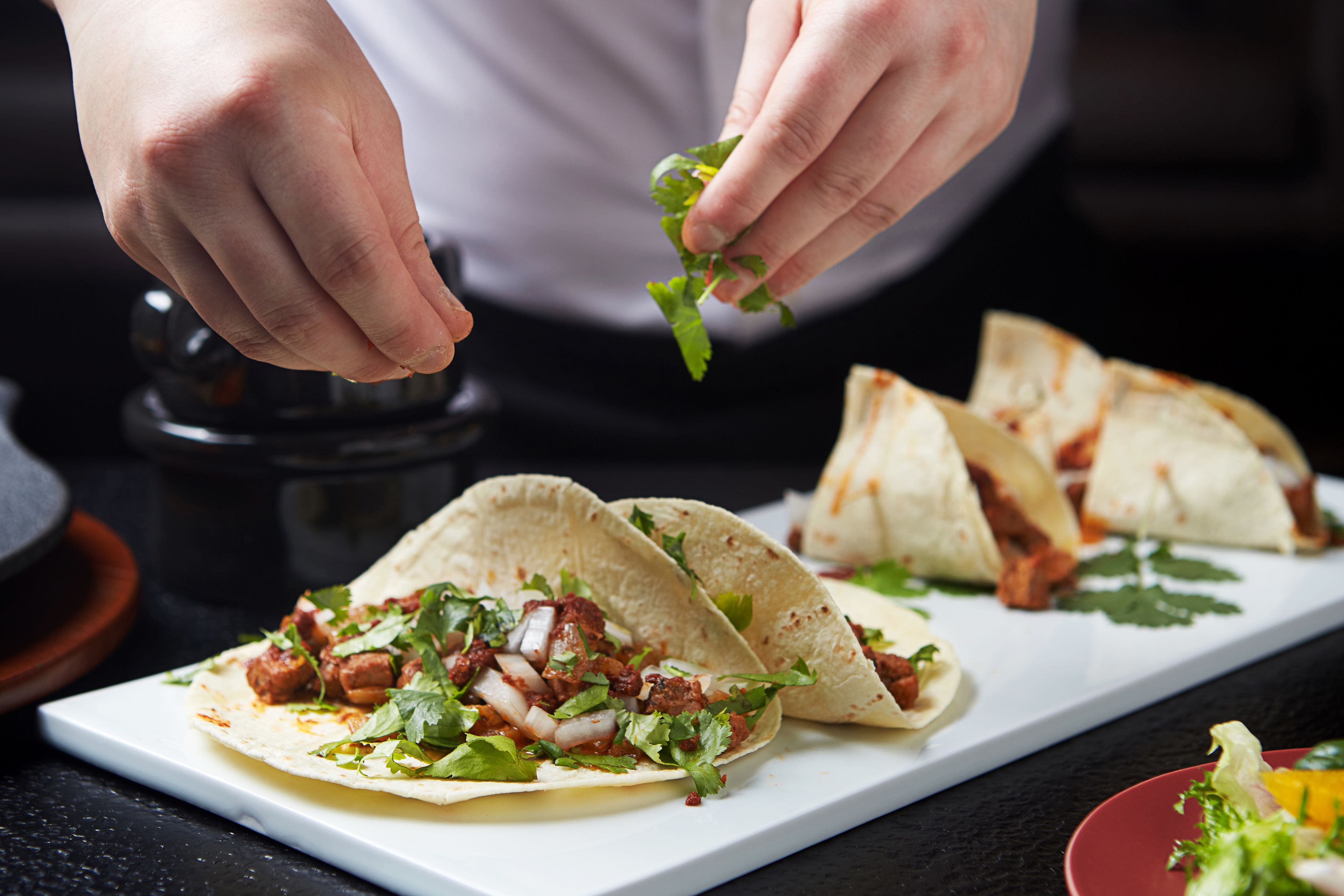 A person preparing tacos on a white plate with a dark background.