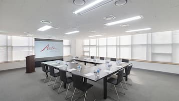 Un salón de conferencias con una pantalla grande que muestra 'Asti Hotel Business Center', una mesa larga con sillas y un atril.
