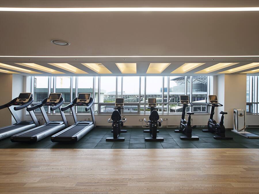 A gym with treadmills and stationary bikes arranged in rows, large windows providing natural light, and a wooden floor.