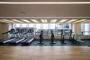 A gym with treadmills and stationary bikes arranged in rows, large windows providing natural light, and a wooden floor.