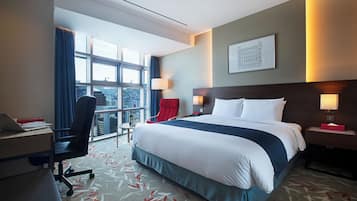 A hotel room with a large bed, a desk with a laptop, a chair, and a window with a city view.