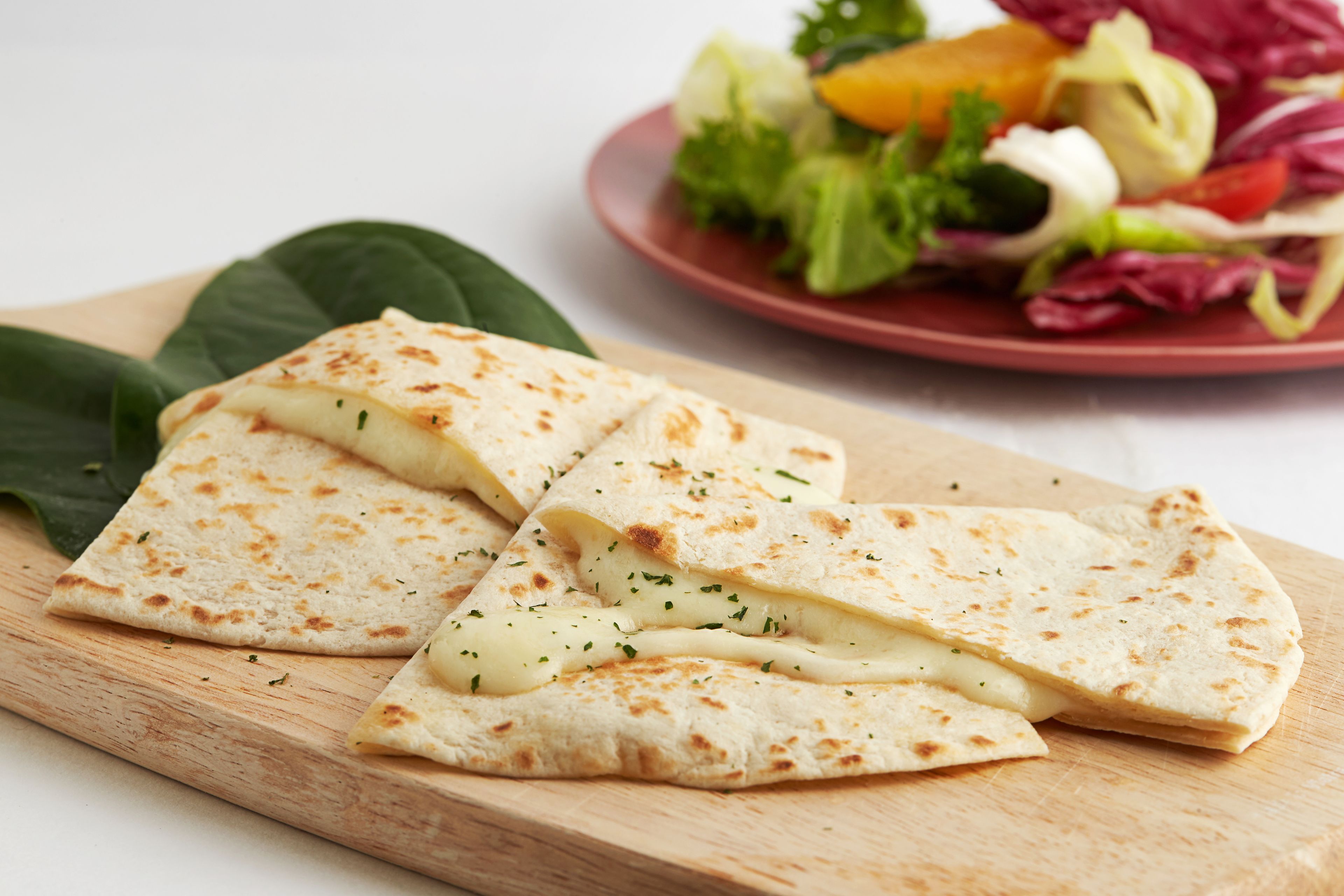 Une planche à découper en bois sur laquelle est posé un pain plat plié, et en arrière-plan, une assiette de salade composée et de légumes.