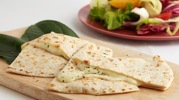 A wooden cutting board with a folded flatbread and a plate of mixed greens and vegetables in the background.